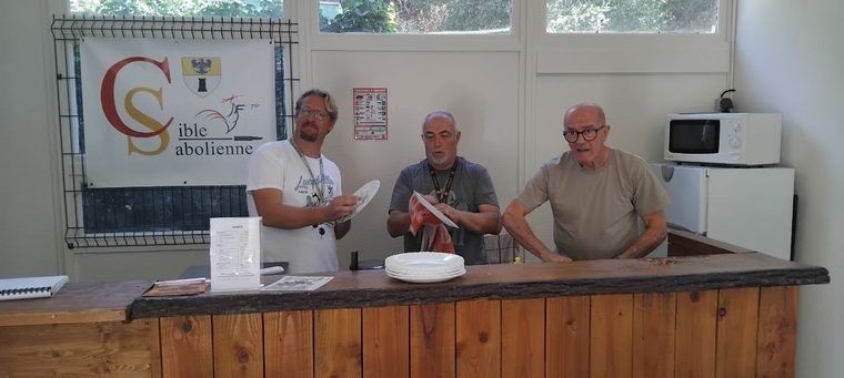 Trois bénévoles essuient la vaisselle derrière le comptoir du club house, sous la banderole de La Cible Sabolienne